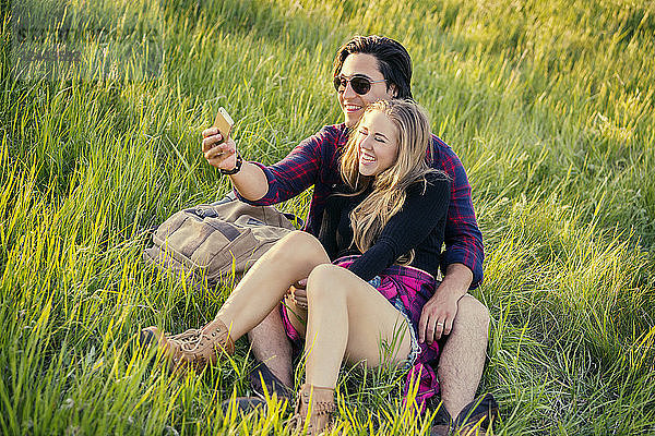 Ein junges Paar sitzt im Gras in einem Park und stellt sich mit seinem Handy für ein Selbstporträt auf  Edmonton  Alberta  Kanada