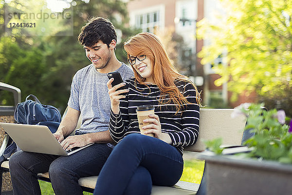 Ein junger Mann und eine junge Frau sitzen zusammen auf einer Bank auf dem Universitätscampus  benutzen einen Laptop und überprüfen soziale Medien auf einem Smartphone  während sie Kaffee trinken  Edmonton  Alberta  Kanada