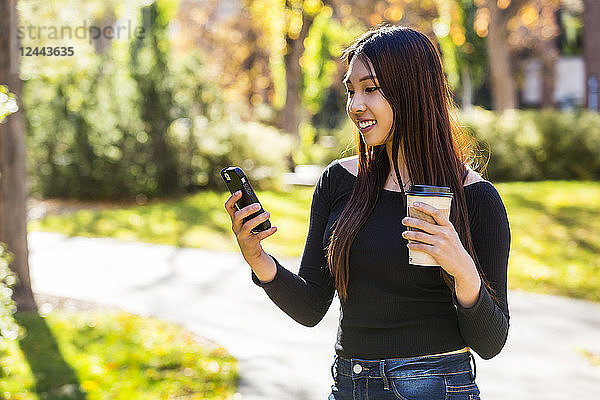 Eine junge chinesische internationale Studentin steht mit einer Kaffeetasse in der Hand und benutzt ihr Smartphone auf dem Universitätscampus  Edmonton  Alberta  Kanada