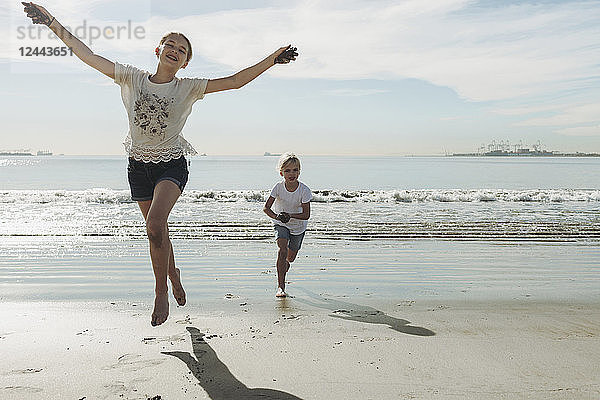 Zwei Schwestern rennen und spielen am Strand  die Älteste vorne nimmt Blickkontakt mit der Kamera auf und springt in die Luft; Long Beach  Kalifornien  Vereinigte Staaten von Amerika