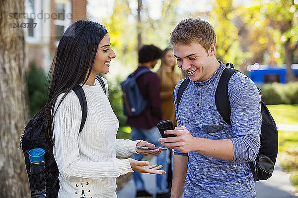 Ein männlicher und ein weiblicher Universitätsstudent stehen auf einem Weg und schauen gemeinsam auf ein Smartphone  lachen und unterhalten sich mit einer kleinen Gruppe von Kommilitonen im Hintergrund  Edmonton  Alberta  Kanada
