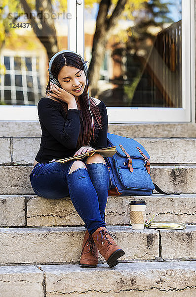 Eine junge chinesische Studentin sitzt auf einer Treppe und hört mit geschlossenen Augen über Kopfhörer Musik  Edmonton  Alberta  Kanada