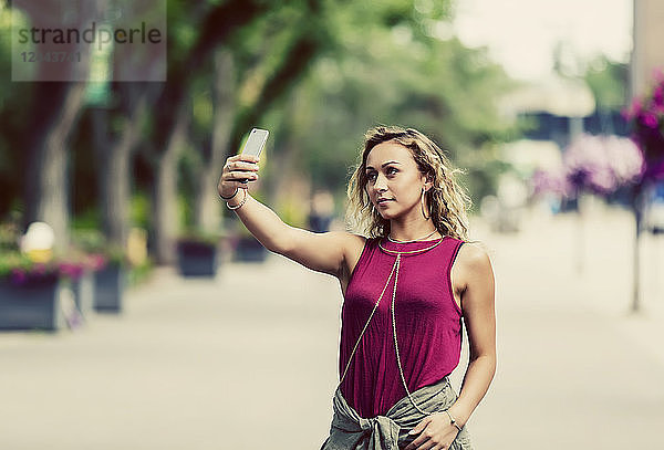 Eine konzeptionelle Aufnahme einer jungen Frau  die ein Selbstporträt mit ihrem Smartphone aufnimmt  während sie allein auf der Straße in der Nähe eines Universitätsgeländes unterwegs ist  Edmonton  Alberta  Kanada