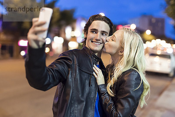Ein junges Paar macht ein Selbstporträt mit einem Smartphone  während die Frau den Mann in der Abenddämmerung auf die Wange küsst  Edmonton  Alberta  Kanada