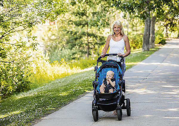 Eine hübsche junge Mutter geht mit ihrer kleinen Tochter im Kinderwagen in einem Park mit einem See an einem warmen sonnigen Tag spazieren  Edmonton  Alberta  Kanada