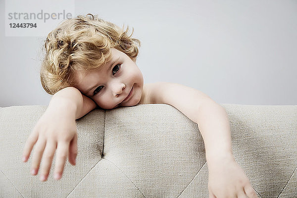 Ein niedlicher kleiner Junge mit langem  lockigem  blondem Haar  der sich an die Rückenlehne einer Couch lehnt und müde aussieht; Langley  British Columbia  Kanada