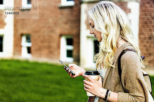Eine schöne junge Frau mit langen blonden Haaren hält eine Kaffeetasse und schreibt eine SMS auf ihrem Smartphone  während sie über einen Universitätscampus geht  Edmonton  Alberta  Kanada