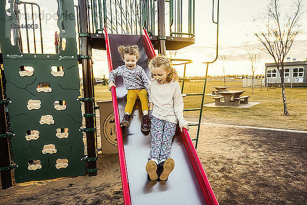 Zwei junge Schwestern rutschen an einem warmen Herbstabend bei Sonnenuntergang auf einem Spielplatz hinunter; Edmonton  Alberta  Kanada