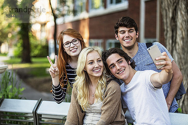 Eine Gruppe von vier Freunden sitzt auf einer Bank und macht ein Selbstporträt mit einem Smartphone auf einem Universitätsgelände  Edmonton  Alberta  Kanada