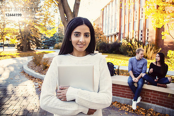 Eine junge Universitätsstudentin libanesischer Abstammung posiert mit ihrem Laptop in der Hand  während sich ihre Freunde im Hintergrund unterhalten  Edmonton  Alberta  Kanada