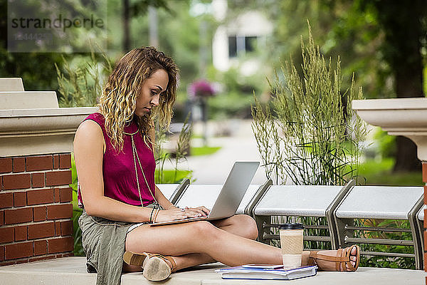 Eine hübsche junge Studentin sitzt an ihrem Laptop auf einer Parkbank draußen auf dem Campus  Edmonton  Alberta  Kanada