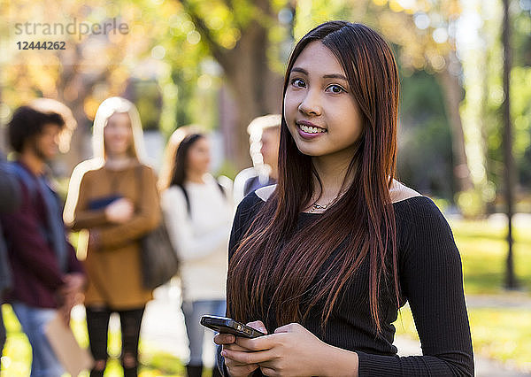 Porträt einer jungen internationalen Universitätsstudentin  die ein Smartphone in der Hand hält  mit ihren Freunden im Hintergrund  Edmonton  Alberta  Kanada