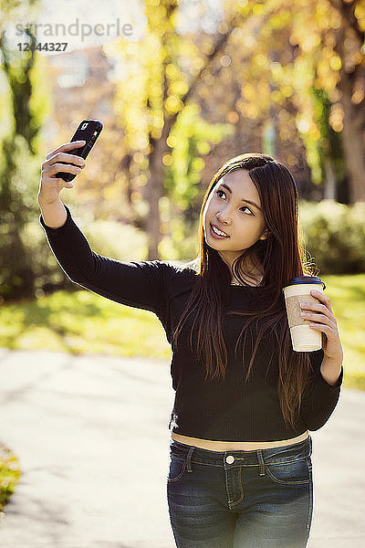 Eine junge internationale Universitätsstudentin hält eine Kaffeetasse in der Hand und posiert für ein Selbstporträt mit ihrem Smartphone  Edmonton  Alberta  Kanada