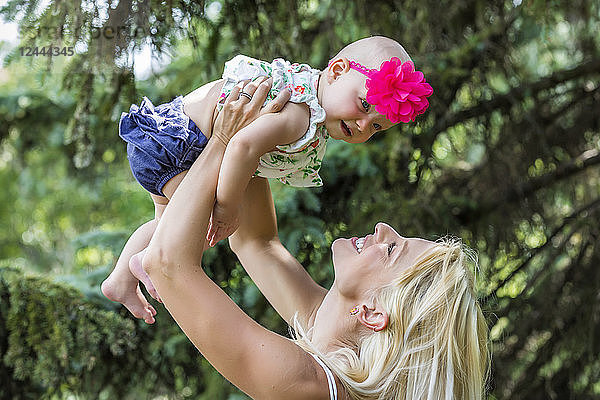 Eine schöne junge Mutter mit langen blonden Haaren genießt die Zeit mit ihrer süßen kleinen Tochter und wirft sie an einem Sommertag in einem Stadtpark in die Luft  Edmonton  Alberta  Kanada