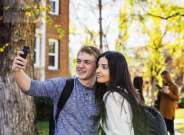 Ein Studentenpaar posiert für ein Selbstporträt mit einem Smartphone und einer kleinen Gruppe von Freunden im Hintergrund  Edmonton  Alberta  Kanada