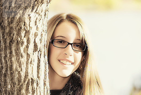 Porträt eines jungen Mädchens mit Brille  das im Herbst hinter einem Baum hervorschaut  Edmonton  Alberta  Kanada