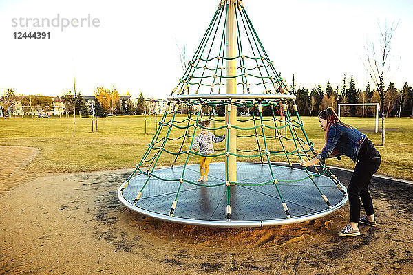 Eine junge Mutter dreht ihre Tochter  während sie auf einem Spielplatz bei Sonnenuntergang an einem warmen Herbstabend mit einem Kletterseil Karussell fährt; Edmonton  Alberta  Kanada