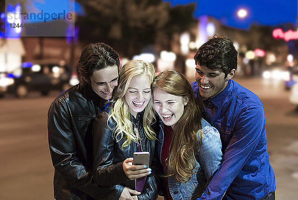 Eine Gruppe von vier Freunden kauert auf einem Bürgersteig zusammen  schaut auf ein Smartphone und lacht  während das Leuchten des Bildschirms ihre Gesichter erhellt  Edmonton  Alberta  Kanada