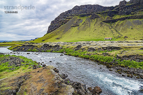 Ein frisch aussehender blauer Fluss fließt am Rande eines Bauernhofs entlang  mit vulkanischen Bergen im Hintergrund  Island