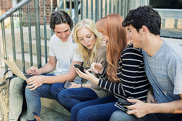 Vier Studenten  die in einer Reihe auf einer Stufe sitzen  ihre Technologie auf dem Universitätscampus nutzen und zusammen lachen  Edmonton  Alberta  Kanada