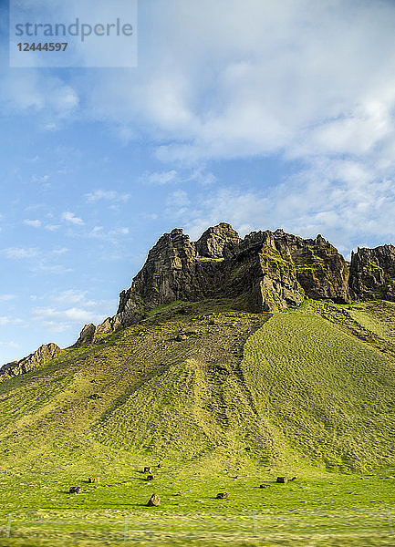 Ein schroffer  felsiger Gipfel  der sich wie ein Monument von den grünen Hügeln und dem blauen Himmel abhebt - ein häufiger Anblick auf einer Reise durch Island  Island