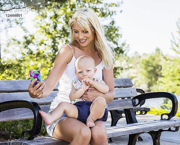 Eine hübsche junge Mutter mit ihrer kleinen Tochter sitzt auf einer Parkbank und versucht  ein Selbstporträt zu machen  während sie an einem warmen Sommertag im Freien Zeit miteinander verbringen  Edmonton  Alberta  Kanada
