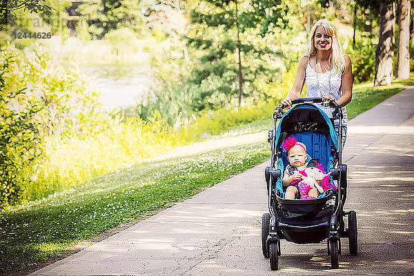 Eine hübsche junge Mutter geht mit ihrer kleinen Tochter im Kinderwagen in einem Park mit einem See an einem warmen sonnigen Tag spazieren  Edmonton  Alberta  Kanada