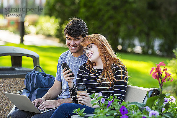 Ein junger Mann und eine junge Frau sitzen zusammen auf einer Bank auf dem Universitätscampus  benutzen einen Laptop und überprüfen soziale Medien auf einem Smartphone  während sie Kaffee trinken  Edmonton  Alberta  Kanada