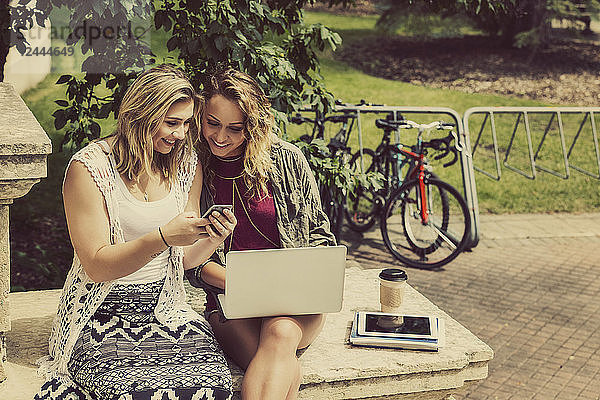 Zwei Studentinnen sitzen zusammen und benutzen ihr Smartphone und ihren Laptop  während sie auf den Stufen eines Universitätsgebäudes sitzen  Edmonton  Alberta  Kanada