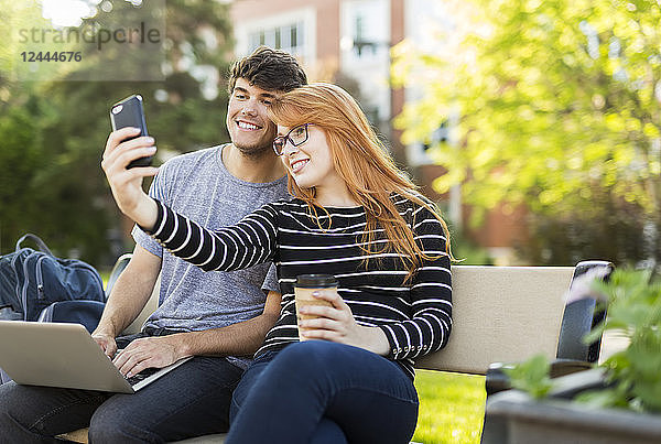 Ein junger Mann und eine junge Frau sitzen zusammen auf einer Bank auf dem Universitätscampus und machen ein Selbstporträt mit einem Smartphone  während sie Kaffee trinken  Edmonton  Alberta  Kanada