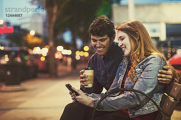 Ein junges Paar sitzt in der Abenddämmerung auf einer Bank an einer beliebten Straße und schaut auf sein Smartphone  Edmonton  Alberta  Kanada