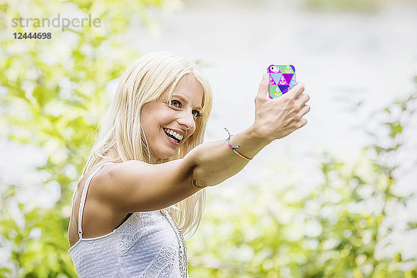 Eine schöne junge Frau  die ein Selbstporträt macht  während sie ihre Zeit in einem Park mit einem künstlichen See genießt  Edmonton  Alberta  Kanada