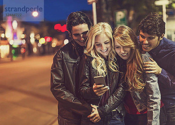 Eine Gruppe von vier Freunden kauert auf einem Bürgersteig zusammen und schaut auf ein Smartphone  während das Leuchten des Bildschirms ihre Gesichter erhellt  Edmonton  Alberta  Kanada