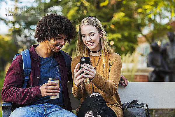 Junges Studentenpaar  das auf einer Bank auf dem Campus sitzt und mit einem Smartphone soziale Medien nutzt  Edmonton  Alberta  Kanada