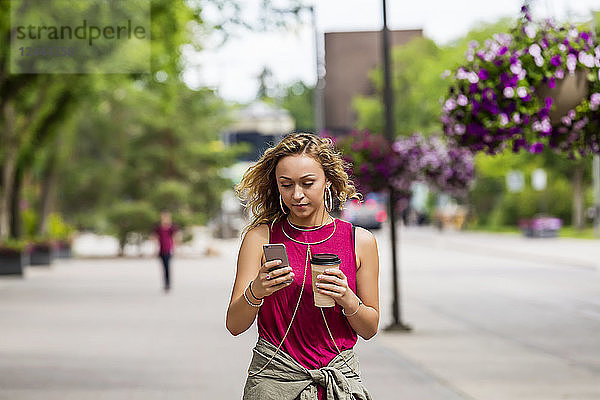 Eine junge Frau geht eine Straße in der Nähe eines Universitätsgeländes entlang und schreibt eine SMS auf ihrem Smartphone  Edmonton  Alberta  Kanada