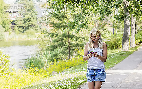 Eine schöne junge Frau  die an einem warmen sonnigen Tag durch einen Park an einem See spaziert und eine SMS auf ihrem Smartphone schreibt  Edmonton  Alberta  Kanada