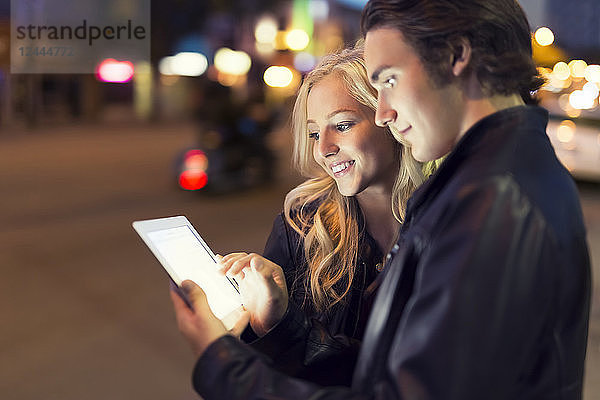 Ein junges Paar benutzt ein Tablet auf einer Straße in der Abenddämmerung  wobei der leuchtende Bildschirm ihre Gesichter erhellt  Edmonton  Alberta  Kanada