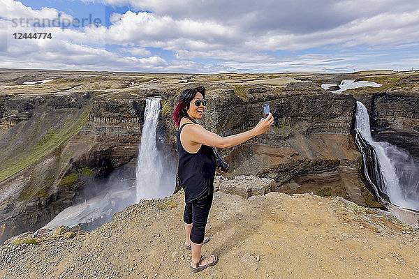 Eine junge asiatische Wanderin posiert für ein Selbstporträt am Rande des Haifoss-Wasserfalltals  einem beliebten Wanderziel in Island  Island