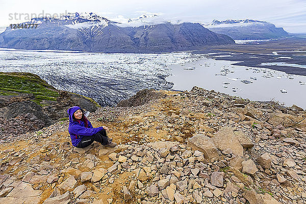 Eine Wanderin in warmer Kleidung posiert auf einem Berggipfel mit Blick auf den Gletschersee und das darunter liegende Tal im Vatnajokull-Nationalpark  Island