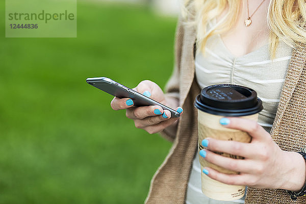 Nahaufnahme der Hände einer jungen Frau mit blauem Nagellack  die eine Kaffeetasse hält und eine SMS auf ihrem Smartphone schreibt  Edmonton  Alberta  Kanada