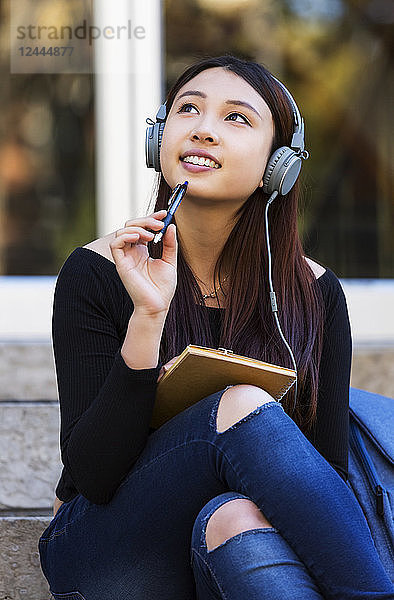 Eine junge chinesische Studentin sitzt auf der Treppe und hört Musik über Kopfhörer. Sie schaut nach oben  während sie träumt und sich einen Stift ans Kinn hält  Edmonton  Alberta  Kanada