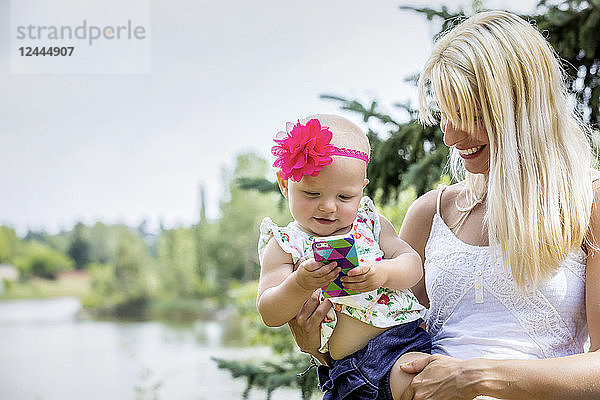 Eine hübsche junge Mutter mit langen blonden Haaren genießt die Zeit mit ihrer süßen kleinen Tochter  die sich mit einem Smartphone in einem Stadtpark an einem Sommertag vergnügt  Edmonton  Alberta  Kanada