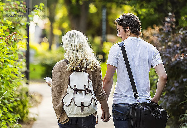 Ein junges Paar geht händchenhaltend einen Weg auf dem Universitätscampus entlang  während die Frau ihr Smartphone auf Nachrichten überprüft  Edmonton  Alberta  Kanada