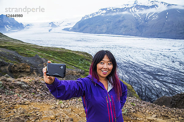 Eine asiatische Wanderin macht ein Selbstporträt auf dem Berggipfel mit dem Gletscher im Hintergrund im Vatnajokull-Nationalpark  Island