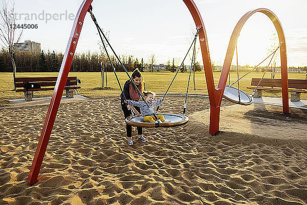 Eine junge Mutter und ihre Tochter spielen auf einer Untertassenschaukel auf einem Spielplatz an einem warmen Herbstabend; Edmonton  Alberta  Kanada