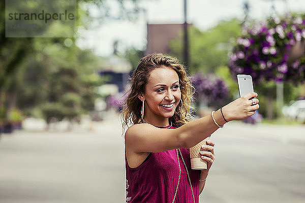 Eine junge  hübsche Frau macht ein Selbstporträt mit einem Handy auf einer Straße in der Nähe eines Universitätsgeländes  Edmonton  Alberta  Kanada