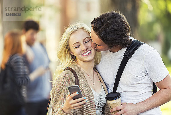 Ein junger Mann küsst eine junge Frau auf die Wange  während sie auf einem Universitätscampus in Edmonton  Alberta  Kanada  soziale Medien auf ihrem Smartphone abruft.