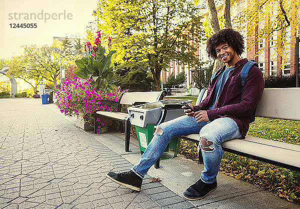 Ein junger afroamerikanischer Student sitzt mit seinem Smartphone auf einer Bank auf dem Universitätscampus  Edmonton  Alberta  Kanada