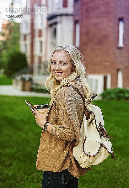 Eine hübsche junge Frau mit langen blonden Haaren hält eine Kaffeetasse in der Hand und schreibt eine SMS auf ihrem Smartphone  während sie auf einem Universitätsgelände spazieren geht und für die Kamera posiert  Edmonton  Alberta  Kanada