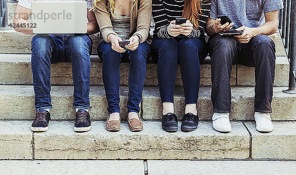 Vier Studenten  die in einer Reihe auf einer Stufe sitzen und ihre Technologie auf dem Universitätscampus nutzen  wobei nur ihre Füße und Beine zu sehen sind  Edmonton  Alberta  Kanada